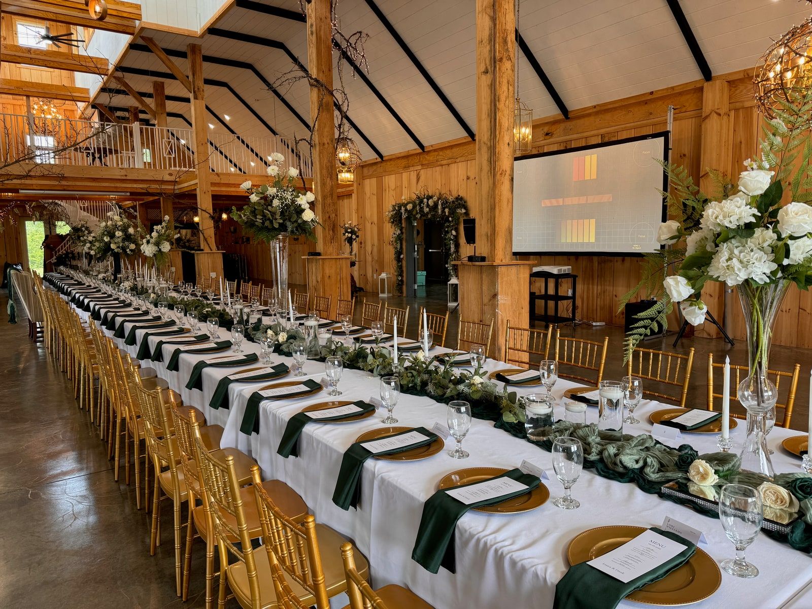 A long banquet table set for a wedding reception inside a two-story barn venue, with a fast-fold projection screen at the front of the room.